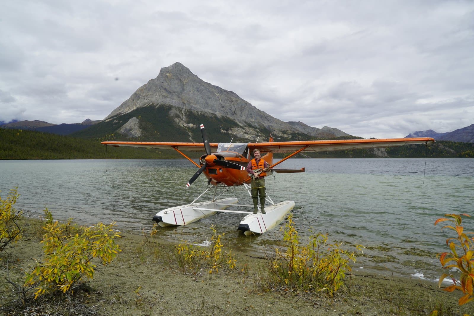 A pilot wearing a life vest stands on the floats of an orange float plane, which is partially beached on the sandy shore of Takahula Lake. A large, rocky mountain rises prominently in the background under a cloudy sky, with forested slopes showing hints of autumn color.