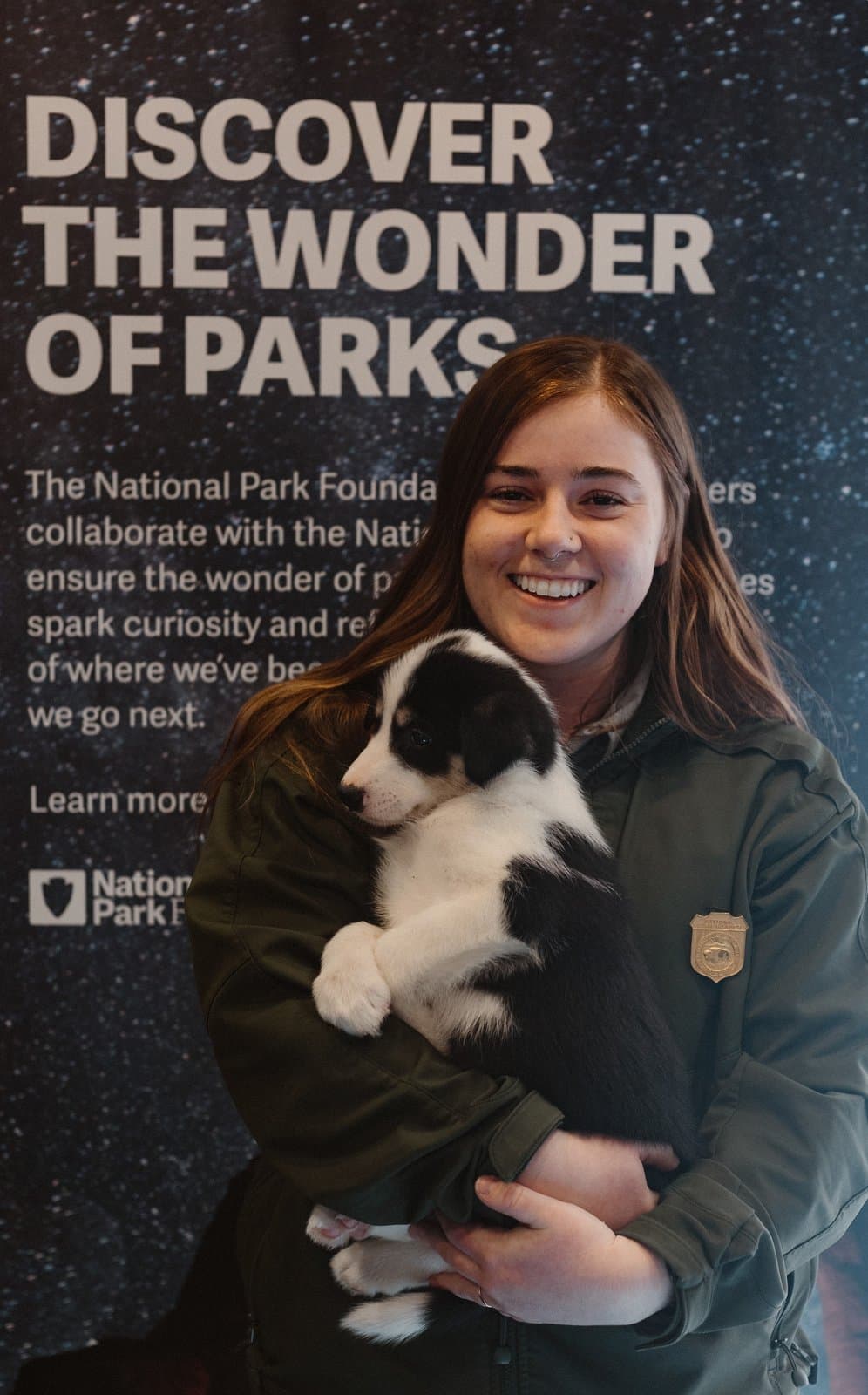 A smiling National Park Service ranger holds a black and white puppy in front of a National Park Foundation display banner.