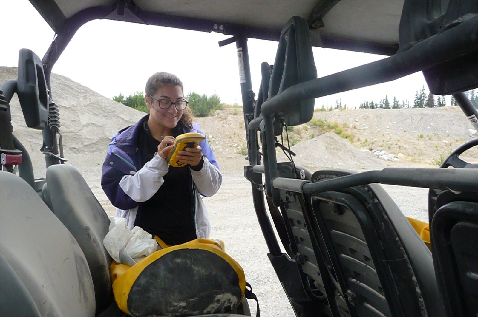 A young woman wearing glasses and a blue and white jacket smiles while using a yellow handheld device and stylus, likely collecting data. She is standing next to a vehicle in an outdoor area with large piles of gravel and distant trees under a cloudy sky.