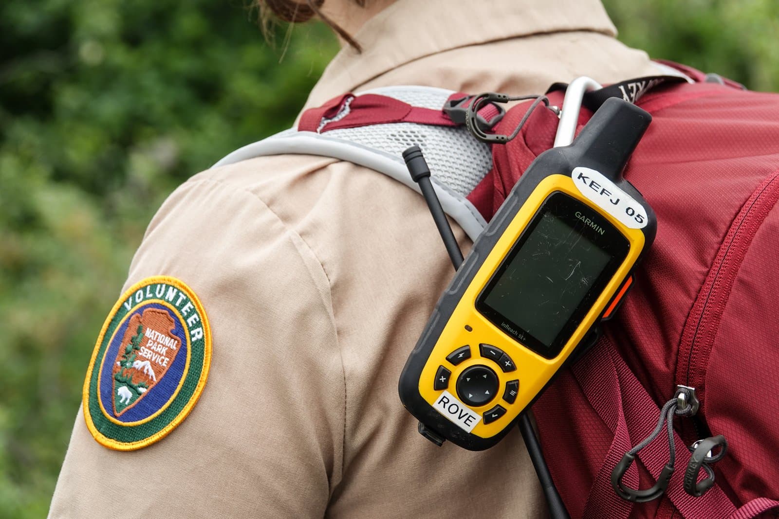 A National Park Service volunteer wears a red backpack equipped with a yellow Garmin inReach satellite communicator while hiking outdoors.
