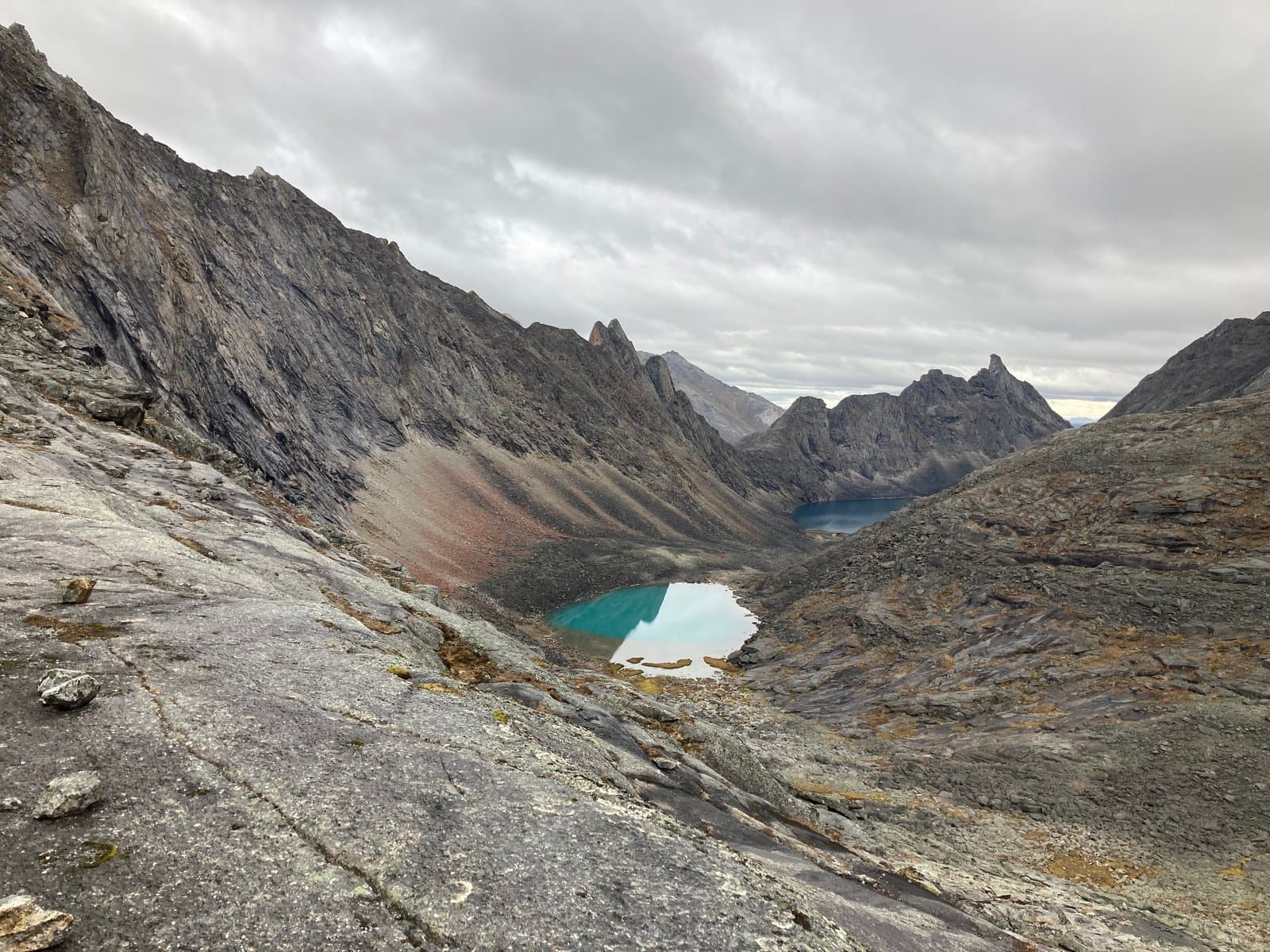 An expansive view of the rugged Arrigetch Valley in Alaska, showcasing two vibrant turquoise alpine lakes nestled among steep, rocky mountains under an overcast sky. The foreground features a rocky slope leading down into the valley.