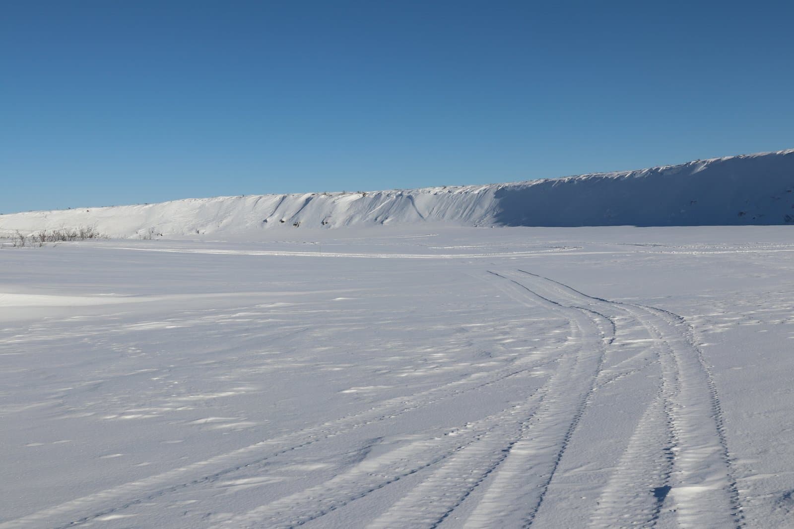 Snowmobile tracks carve a path across the frozen, snow-covered expanse of the Kick River bottom under a clear blue sky.