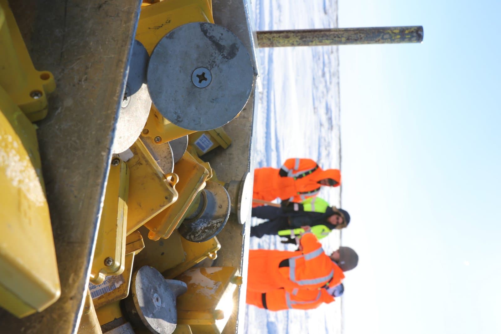 A field crew in high-visibility orange gear works near seismic nodes on a snowy, open landscape in Alaska.