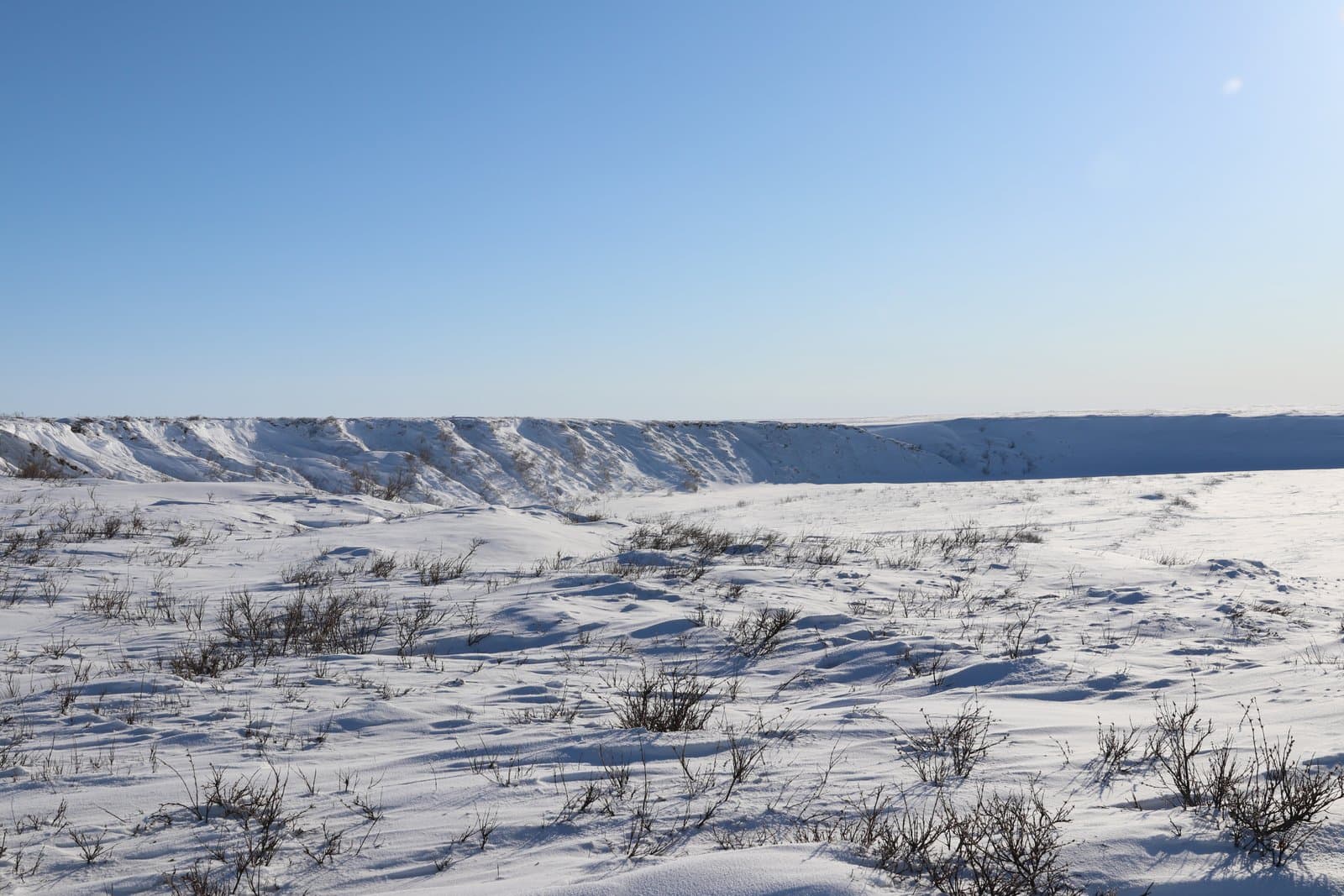 Snow-covered bluffs rise above the frozen, shrub-dotted landscape along the Kick River under a clear, bright blue sky in late March.