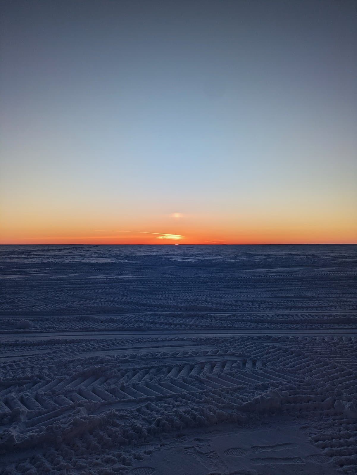 A vibrant spring sunset glows over the vast, snow-covered landscape of the National Petroleum Reserve in Alaska, marked by deep tire tracks.