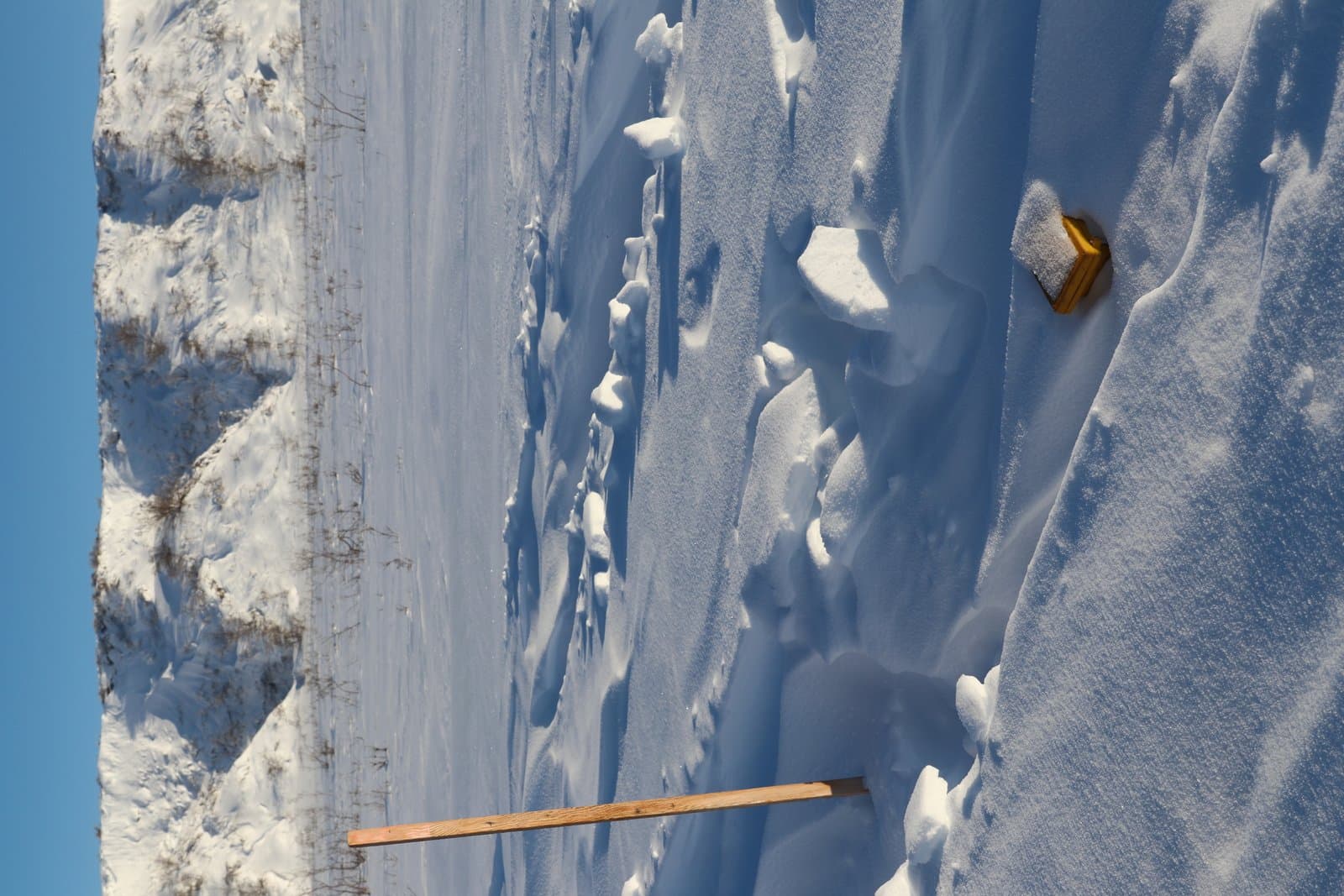 A yellow seismic node and a wooden marker stake sit partially buried in deep, wind-swept snow against a snowy hillside.