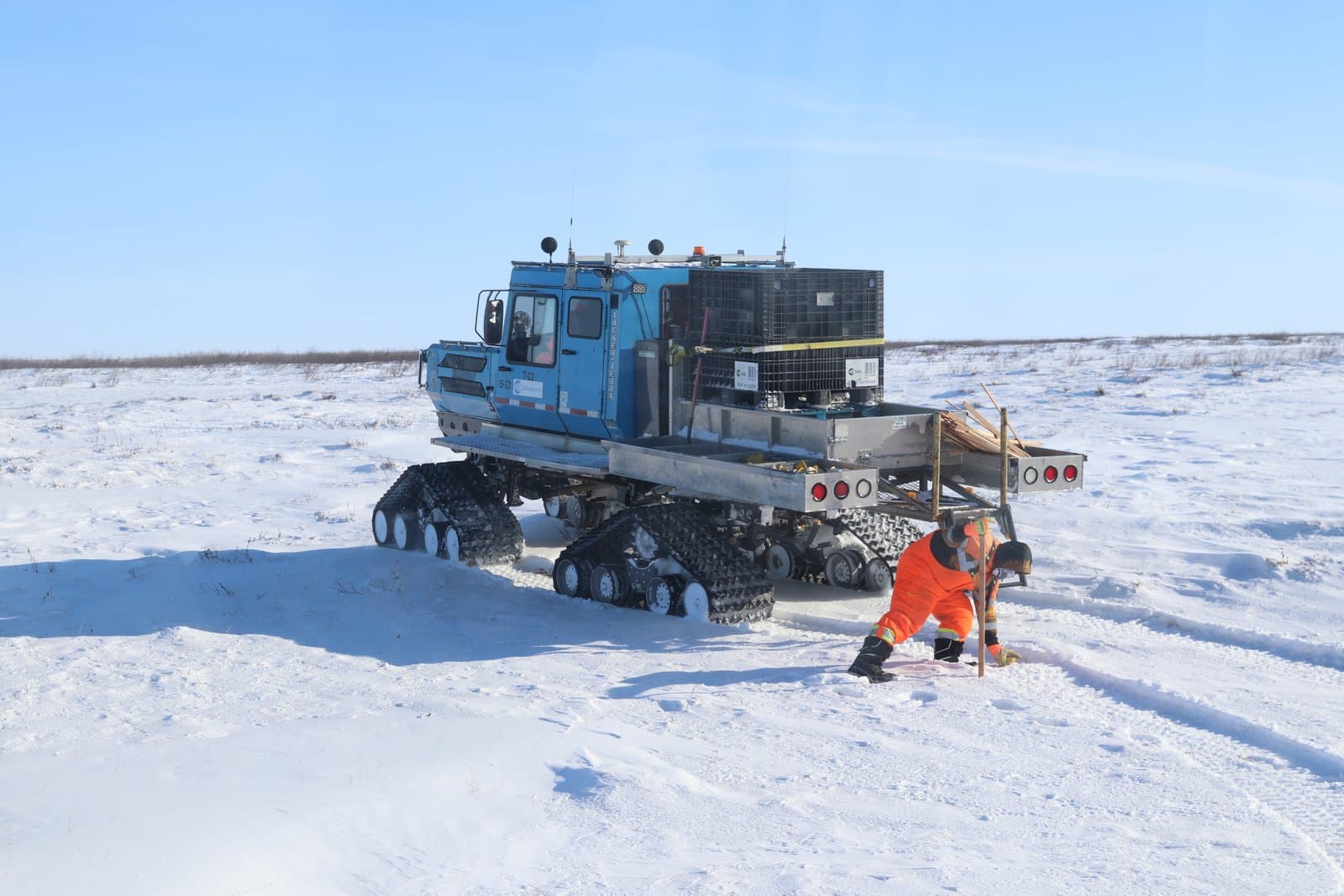 A worker in high-visibility orange gear places a seismic node into the snow next to a blue Tucker-Terra tracked vehicle on the tundra.