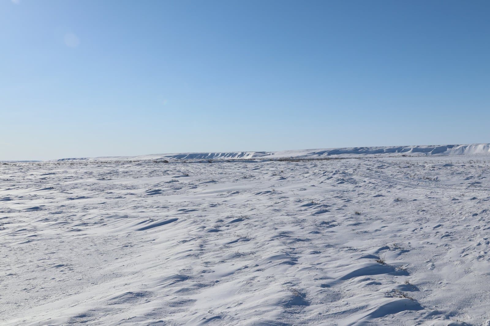 Snow-covered tundra stretches toward the distant, wind-swept Colville River Bluffs under a clear, bright blue sky in rural Alaska.