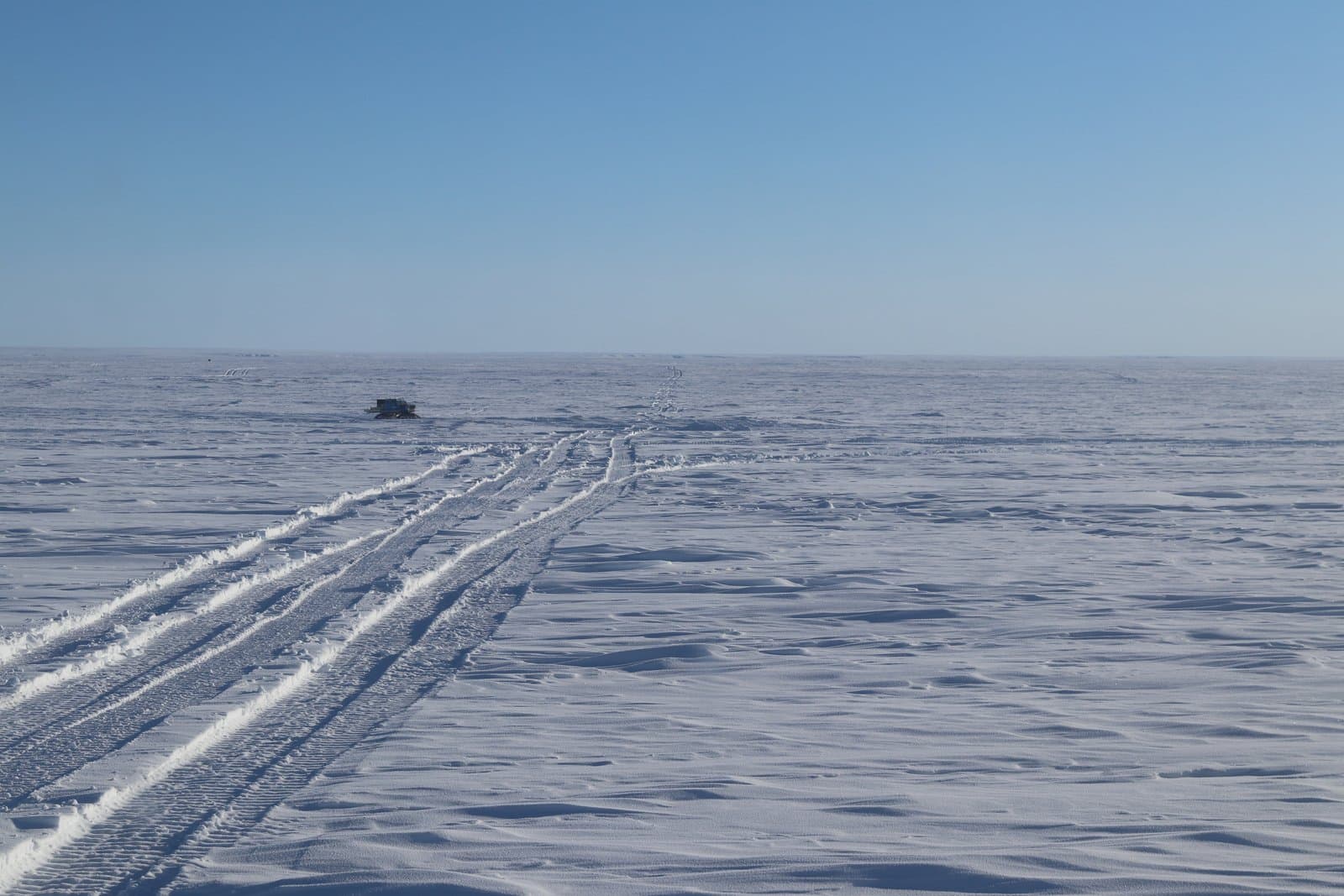 A Tucker snowcat traverses the vast, snow-covered tundra, leaving deep tracks across the flat, frozen landscape under a clear blue sky.