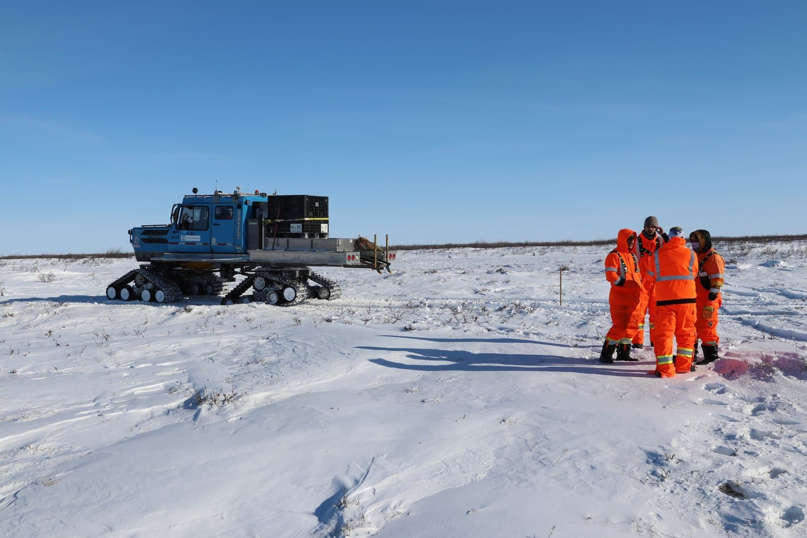 A crew in high-visibility orange gear discusses their next route while standing near a tracked vehicle on a vast, snow-covered Arctic landscape.
