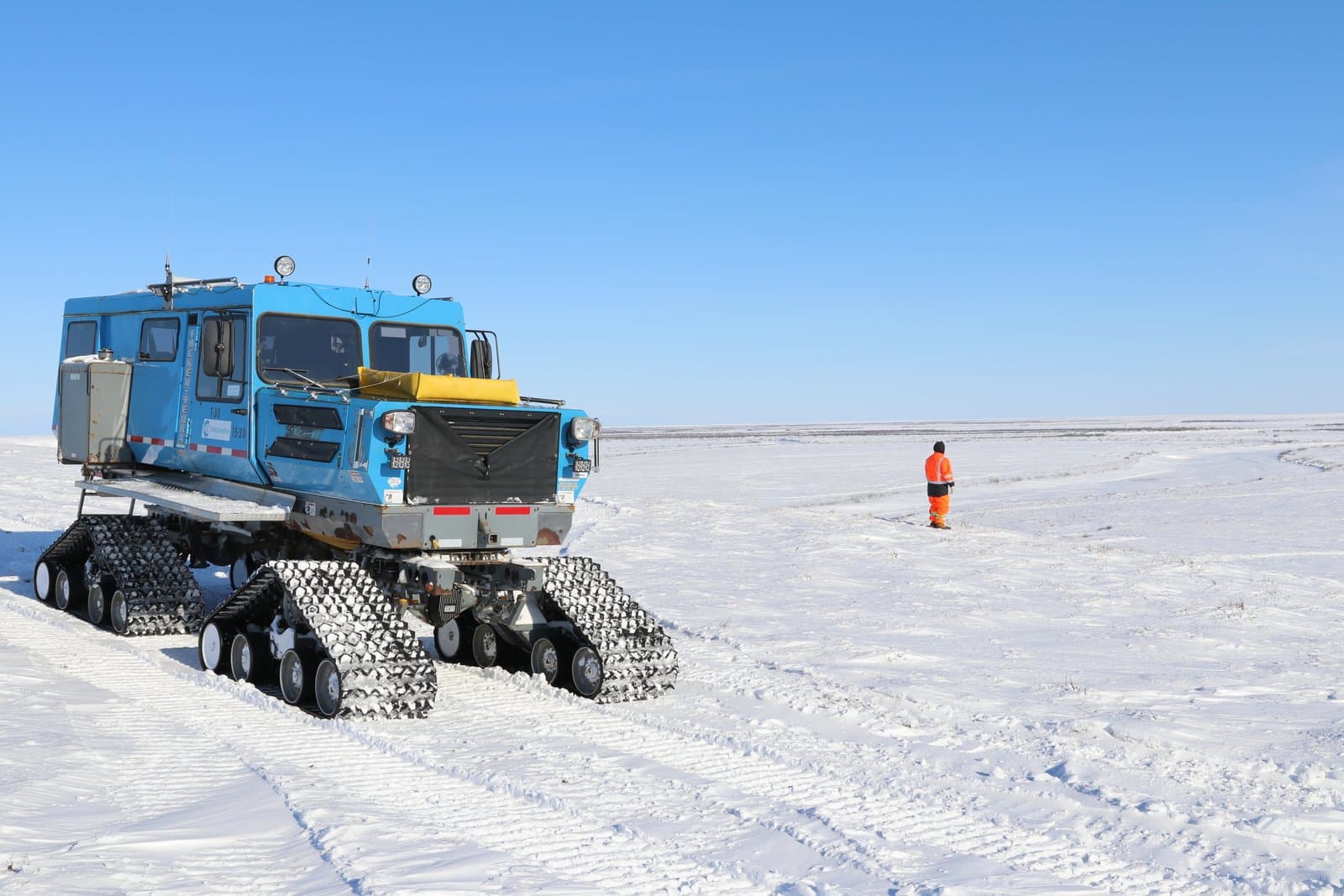 A blue Tucker snow vehicle sits on a snowy bluff overlooking the Colville River while a worker in orange gear stands nearby.