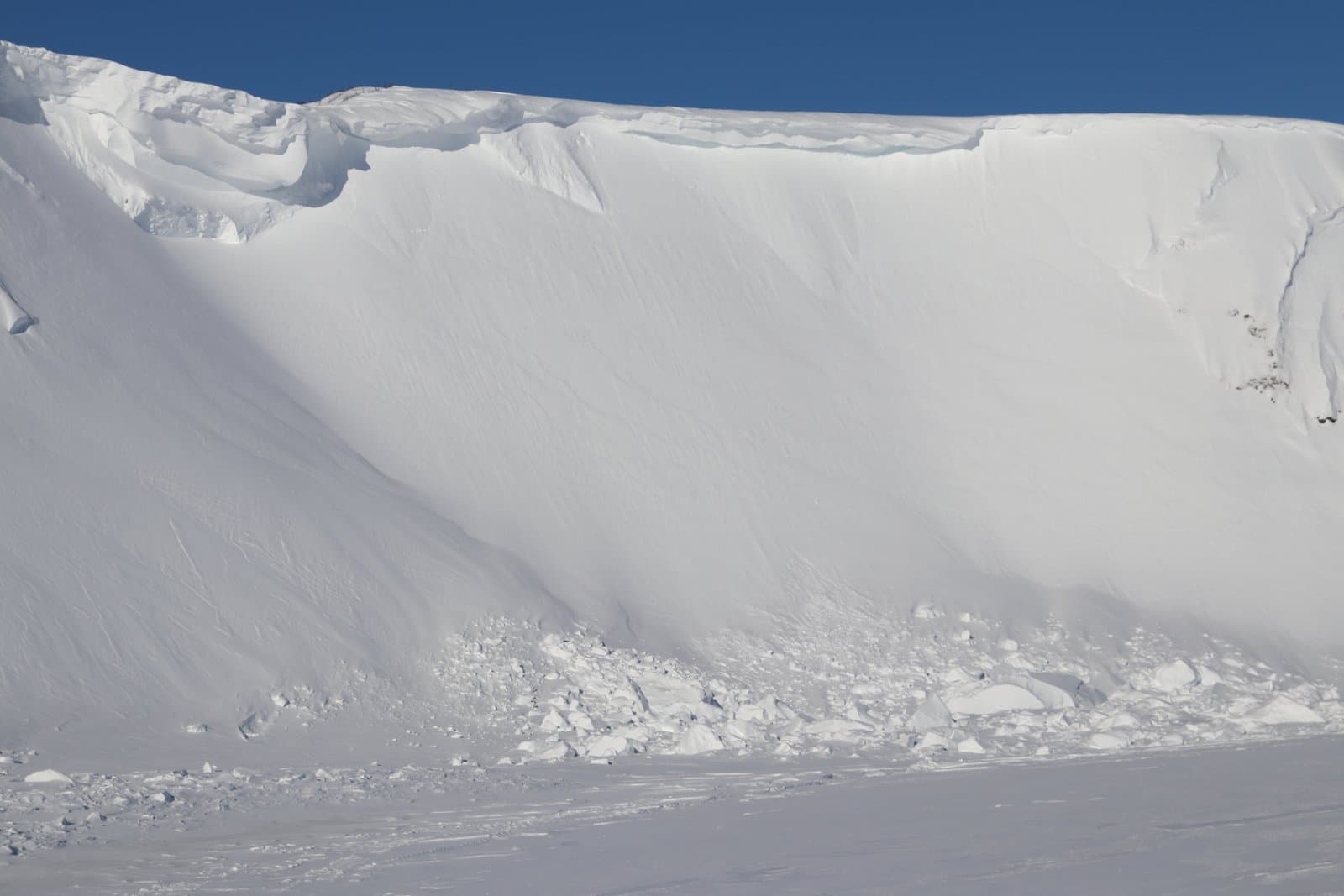 A massive, snow-covered bluff rises above the frozen Colville River, featuring a sharp cornice and debris from a recent snow slide.