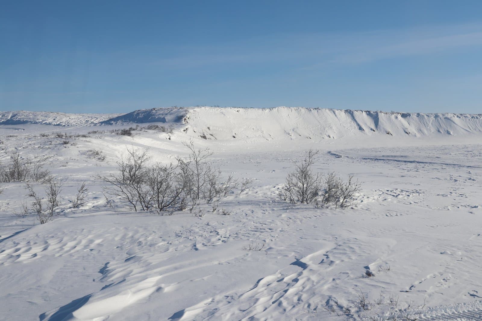 Snow-covered landscape of the Kick River valley featuring frozen terrain, sparse shrubs, and a large snow-drifted embankment under a clear blue sky.