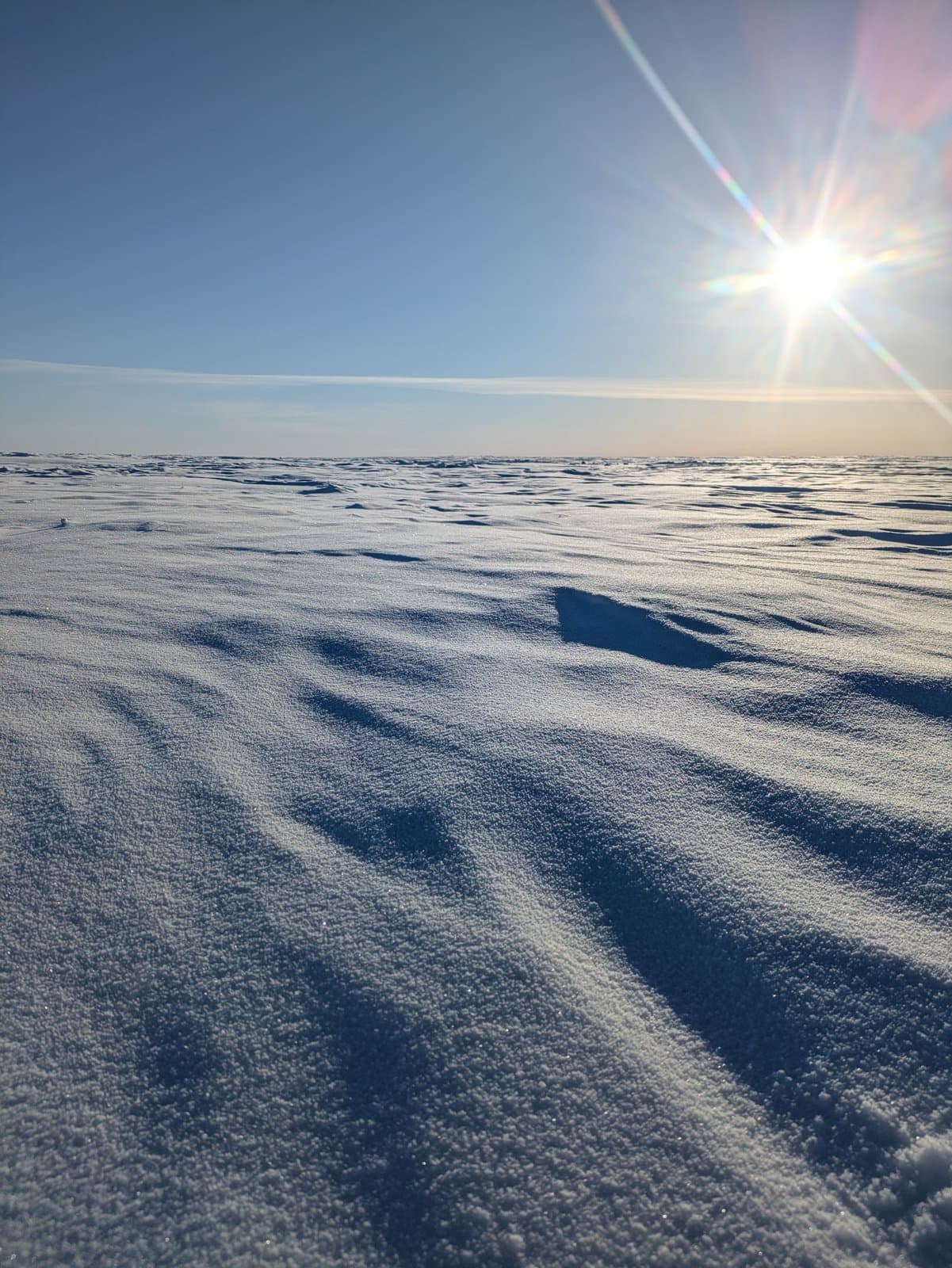 Wind-driven snow patterns stretch across the vast, frozen landscape of the National Petroleum Reserve in Alaska under a bright, low-hanging sun.